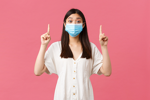 Covid-19, social distancing, virus and lifestyle concept. Surprised asian girl in medical mask looking camera intrigued and asking question about product on top, pointing fingers up, pink background.