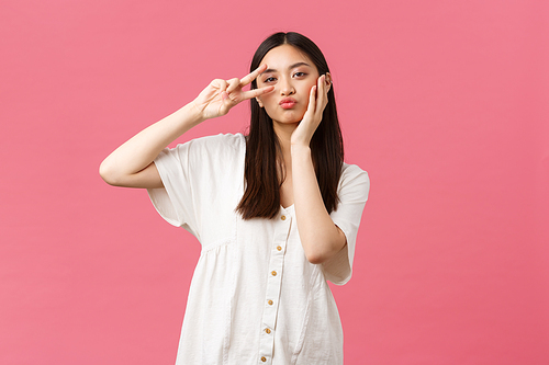 Beauty, people emotions and summer leisure concept. Pretty and stylish asian girl in white dress, posing with kawaii peace sign, touching her cute face, standing pink background.