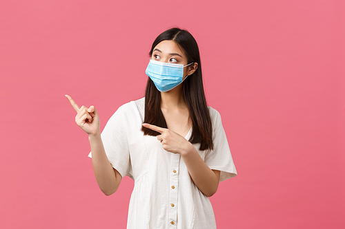 Covid-19, social distancing, virus and lifestyle concept. Curious cute asian girl, store visitor in medical mask asking question about product, pointing fingers left, standing pink background.