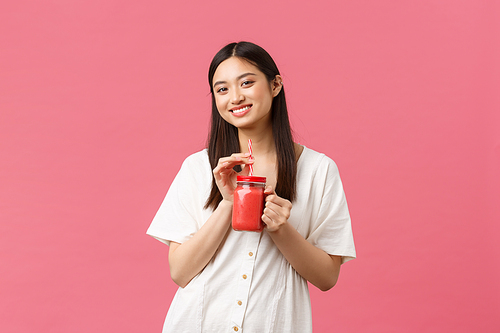 Healthy food, emotions and summer lifestyle concept. Smiling good-looking fit asian girl taking care of body, drinking fresh smoothie from glass and gazing camera satisfied, pink background.