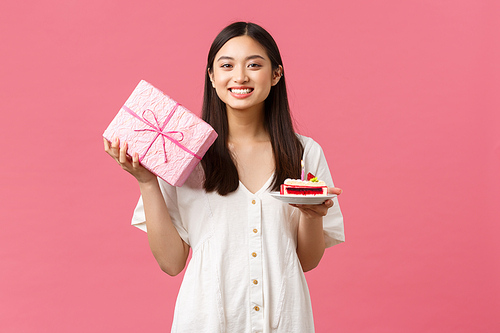 Celebration, party holidays and fun concept. Lovely asian girl in white dress celebrating birthday, holding gift and b-day cake, smiling at friend got her present, pink background.