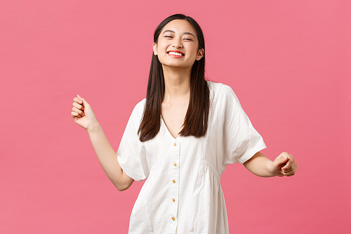 Beauty, people emotions and summer leisure concept. Carefree attractive happy korean girl in white dress dancing and laughing from happiness, standing pink background.