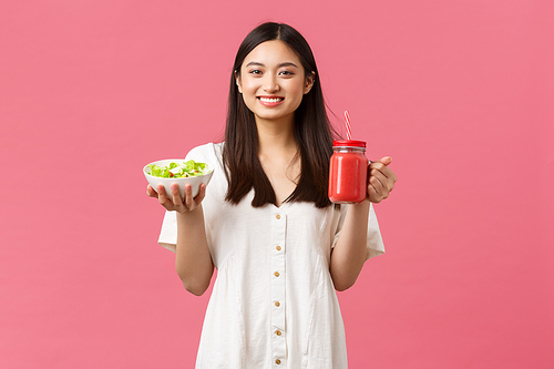 Healthy food, emotions and summer lifestyle concept. Enthusiastic and upbeat cute asian girl full of energy, eating tasty fresh salad and drinking smoothie, smiling at camera happy, pink background.