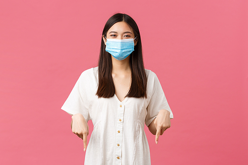 Covid-19, social distancing, virus and lifestyle concept. Delighted cute girl in medical mask and white dress, reading funny sign, pointing fingers down satisfied, smiling over pink background.