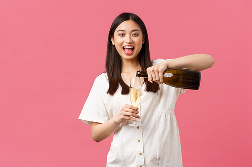 Celebration, party holidays and fun concept. Happy carefree asian woman on vacation, pour champagne in glass and laughing, enjoying weekend or leisure time, standing pink background.