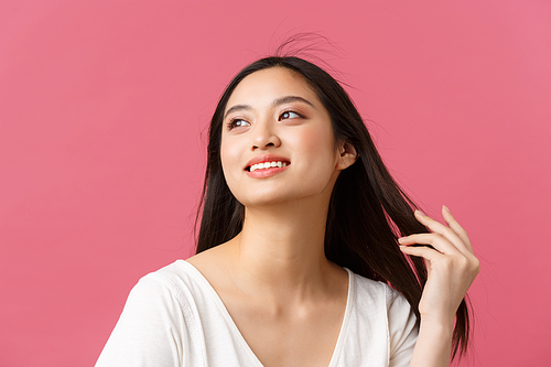 Beauty products advertisement, haircare and women fashion concept. Romantic tender asian woman with beautiful white smile, looking away dreamy as touching hair, standing pink background.