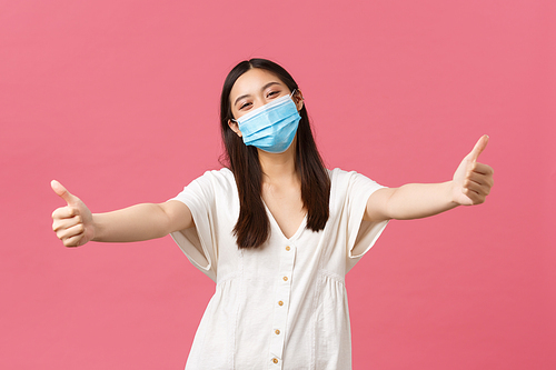 Covid-19, social distancing, virus and lifestyle concept. Friendly happy asian woman in medical mask and summer dress, extend hands to give hug, welcome someone, standing pink background.