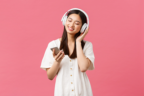 Beauty, people emotions and technology concept. Carefree sensual and pretty asian girl enjoying music in wireless headphones, close eyes and smiling listening favorite song, holding smartphone.