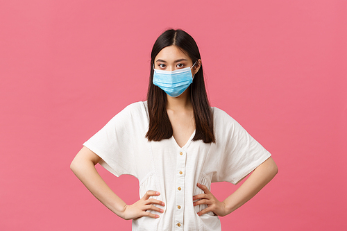 Covid-19, social distancing, virus and lifestyle concept. Serious-looking asian girl in medical mask, hold hands on waist and scolding persong not using hand sanitizer while going out.