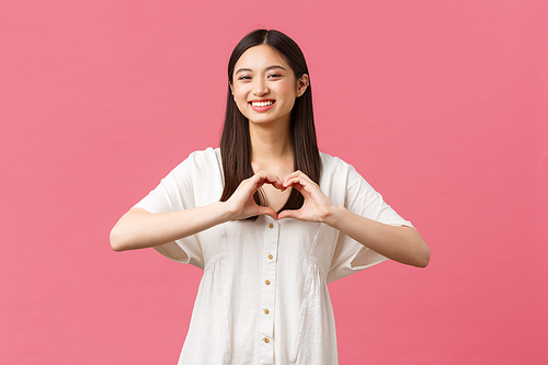 Beauty, people emotions and summer leisure concept. Lovely, romantic asian girl in white dress confess sympathy or love, like someone, showing heart sign and smiling over pink background.