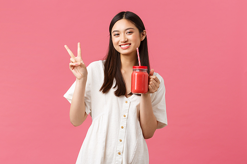 Healthy food, emotions and summer lifestyle concept. Happy good-looking asian girl in white dress, showing kawaii peace sign and drinking smoothie from glass and straw.