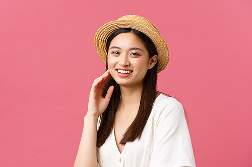 Beauty, people emotions and summer leisure and vacation concept. Happy asian female tourist enjoying weekend, looking camera with broad cheerful smile, wearing straw hat, pink background.