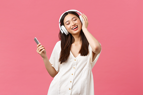 Beauty, people emotions and technology concept. Carefree and pretty asian girl enjoying music in wireless headphones, close eyes and smiling listening favorite song, holding smartphone and dancing.