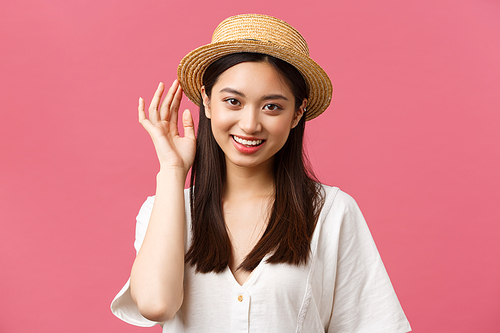 Beauty, people emotions and leisure and vacation concept. Lovely asian woman shopping in store, picking new straw hat, smiling delighted, buying summer outfit over pink background.