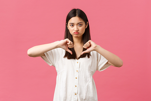 Beauty, people emotions and summer leisure and vacation concept. Judgemental moody and skeptical cute asian girl grimacing disappointed, showing thumbs-down with dislike, pink background.