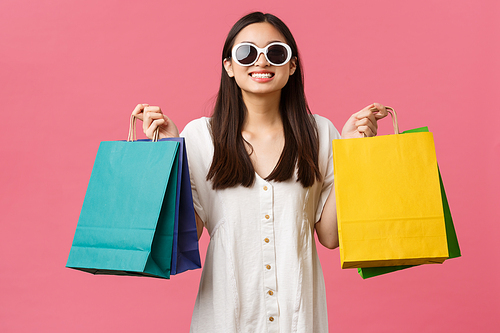 Beauty, people emotions and summer leisure and vacation concept. Carefree happy asian girl on vacation, tourist holding shopping bags and wearing sunglasses, smiling satisfied, pink background.