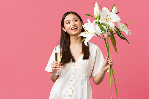 Celebration, party holidays and fun concept. Pretty young woman with glass champagne and bouquet of lilies rejoicing celebrating her birthday, being congratulated with b-day, pink background.