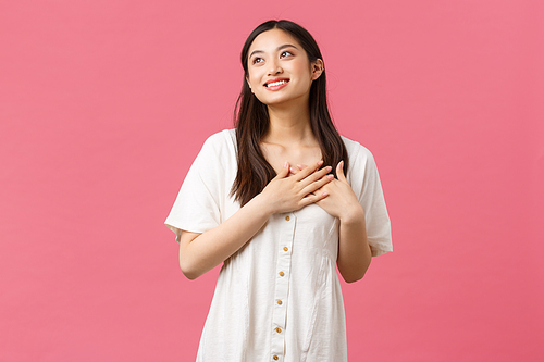 Beauty, people emotions and summer leisure concept. Dreamy and romantic silly asian girlfriend in white dress, looking away imaging perfect date, holding hands on heart touched, pink background.