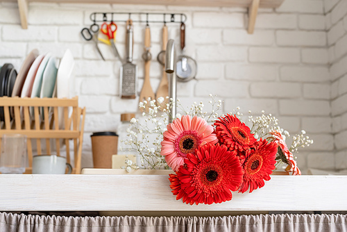 Rustic kitchen interior with white brick wall and white wooden shelves. Red and pink gerbera daisies in a kitchen sink