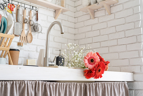 Rustic kitchen interior with white brick wall and white wooden shelves. Red and pink gerbera daisies in a kitchen sink