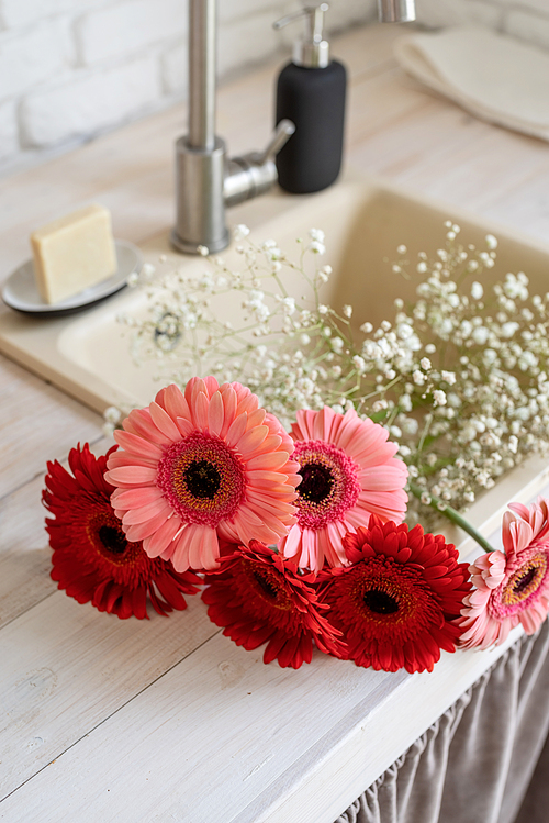 Rustic kitchen interior with white brick wall and white wooden shelves. Red and pink gerbera daisies in a kitchen sink