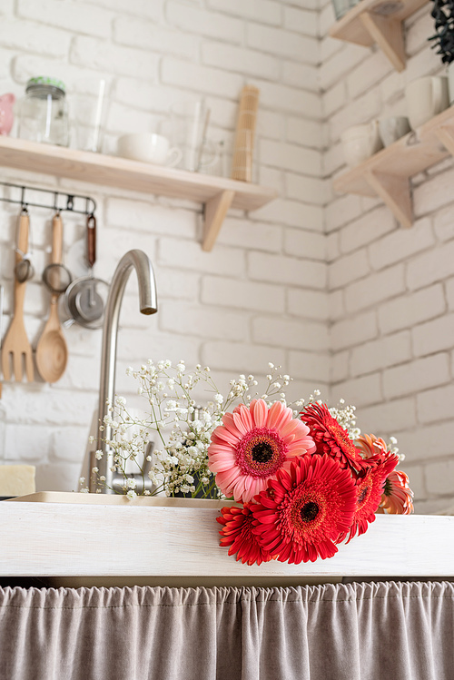 Rustic kitchen interior with white brick wall and white wooden shelves. Red and pink gerbera daisies in a kitchen sink