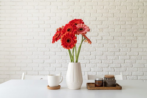 Red and pink gerbera daisies in white vase on kitchen table with candles and cup, minimal style