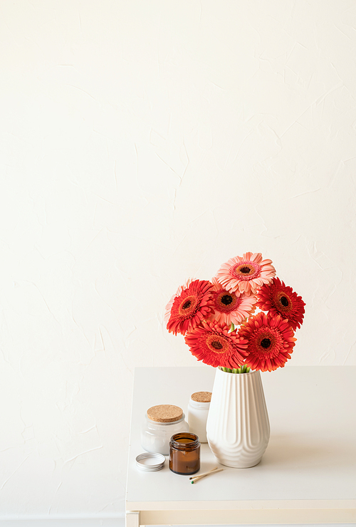 Red and pink gerbera daisies in white vase on kitchen table with candles and cup, minimal style