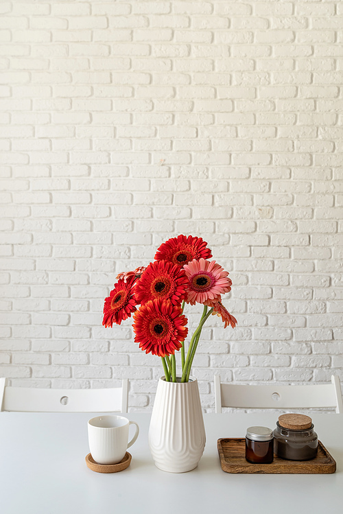 Red and pink gerbera daisies in white vase on kitchen table with candles and cup, minimal style