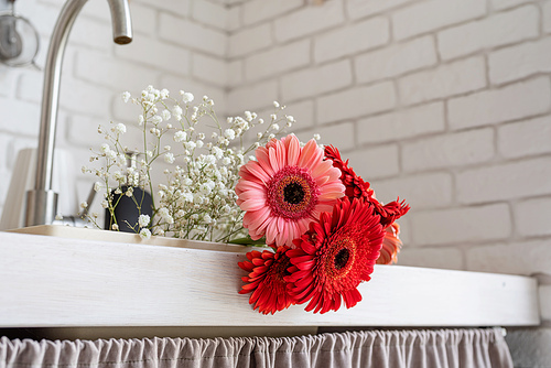 Rustic kitchen interior with white brick wall and white wooden shelves. Red and pink gerbera daisies in a kitchen sink