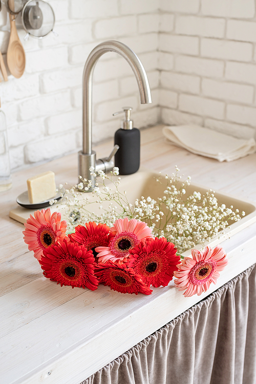 Rustic kitchen interior with white brick wall and white wooden shelves. Red and pink gerbera daisies in a kitchen sink