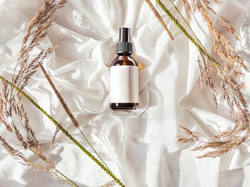 Concept of apothecary cosmetic. Herbs and brown glass bottle of essential oil in still life composition on crumpled white fabric. Sunlight and shadow. Natural cosmetic remedy with white tag.