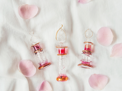 Three graceful bottles for perfume on white crumpled fabric. Top view on pink glass bottles with eastern ornament. Pink rose petals as decoration. Flat lay, still life.