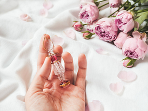 Graceful bottle for perfume or essential oil on white crumpled fabric. Woman holds pink glass bottle with eastern ornament. Pink rose bouquet and petals as decoration.