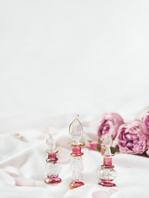 Three graceful bottles for perfume or essential oil on white crumpled fabric. Pink glass bottles with eastern ornament. Pink rose bouquet and petals as decoration. Background with copy space.