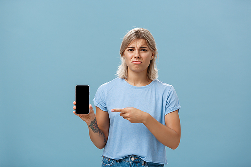 Lifestyle. Unhappy sad attractive silly girl with fair hair tattoos and tanned skin frowning with regret and disappointment poonting at smartphone screen as if being dissatisfied with new girlfriend of ex-lover.