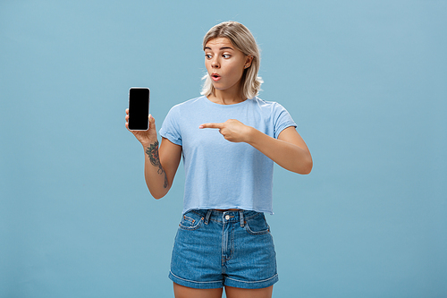 You should download app. Portrait of amazed shocked attractive blonde girl in trendy summer t-shirt holding smartphone gasping from surprise pointing at gadget screen posing over blue background.
