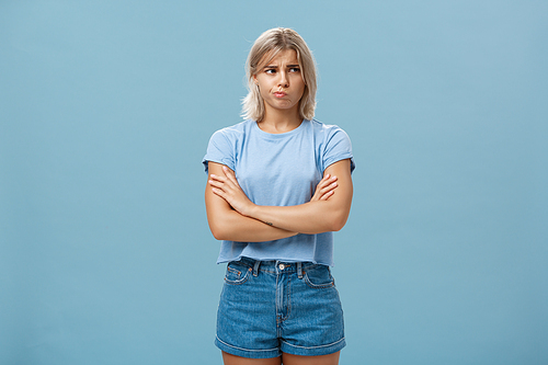 Uncertain troubled and perplexed attractive blond woman with tanned skin holding hands crossed on chest pouting and frowning looking right with worried unsure and doubtful look over blue wall. Feelings and emotions concept