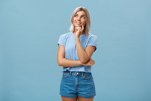 Dreamy thoughtful and creative artistic blonde woman in denim shorts and t-shirt smiling curiously touching lip and gazing at upper left corner feeling nostalgic, thinking over blue wall.