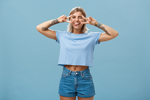 Enjoy life and dream. Portrait of joyful good-looking carefree european female student with blond short haircut winking sticking out tongue entertained and showing peace signs over eyes near blue wall.