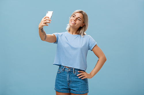 Lifestyle. Stylish cute female gym manager taking selfie with new smartphone posing with broad pleased smile and holding hand on hip making post for followers standing carefree and relaxed over blue wall.