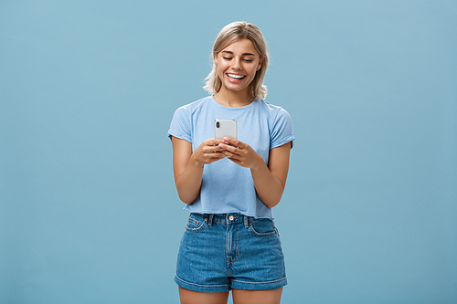Nice to have internet friends. Carefree happy attractive blonde female student with tanned skin and blond hair reading message from smartphone smiling at device screen pleased over blue wall.