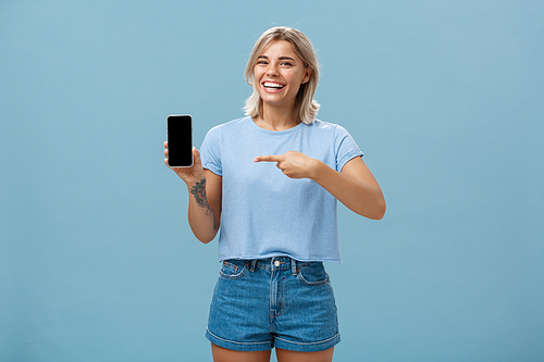Look at this hilarious photo. Entertained attractive happy woman with fair hair in casual t-shirt and denim shorts showing smartphone at camera pointing at device screen smiling broadly over blue wall.
