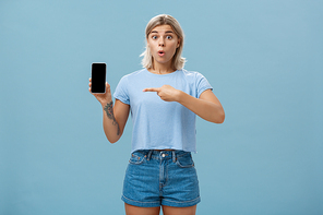 Wow look what found online. Portrait of impressed speechless good-looking young sportswoman in t-shirt and denim shorts saying wow folding lips showing smartphone and pointing at screen over blue wall. Technology concept
