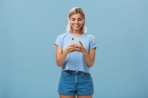 Girl having funny convesation, speaking with friend via video messages in wireless earphones holding smartphone in both hands smiling happily at device screen being entertained and amused. Emotions and technology concept