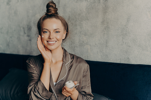 Image of caucasian cheerful woman in silk pajama smiling while gently applying moisturizing face cream isolated over concrete wall background in bedroom. Beauty and skincare concept