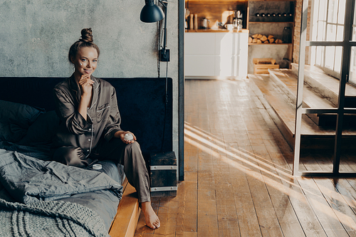 Young beautiful girl sitting on bed in pajama after waking up in stylish apartment bathed in morning sunlight, while holding and applying moisturizing face cream on her skin. Beauty treatment concept