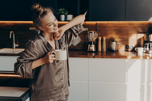 Young pretty relaxed woman in comfy pajama stretching from sleep early in morning while holding cup of coffee in her hand, standing in stylish kitchen interior. Leisure time on average day at home