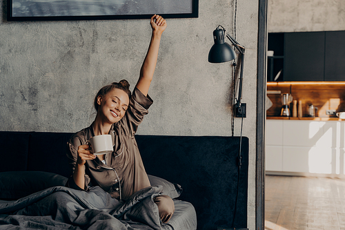 Sleepy relaxed lovely girl in satin pajama trying to wake up from night sleep, sitting in bed with closed eyes while holding her cup of coffee and stretching hand up, enjoying weekend at home