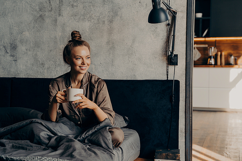 Happy smiling caucasian female in pajama enjoying morning cup of coffee while sitting in bed after waking up before starting her busy work day, relaxing in bedroom of contemporary apartment or hotel